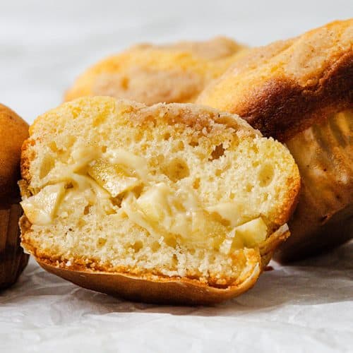 A close-up of four golden Apple Cheesecake Muffins on crinkled white paper, with one muffin cut in half to reveal a creamy filling with apple chunks inside.