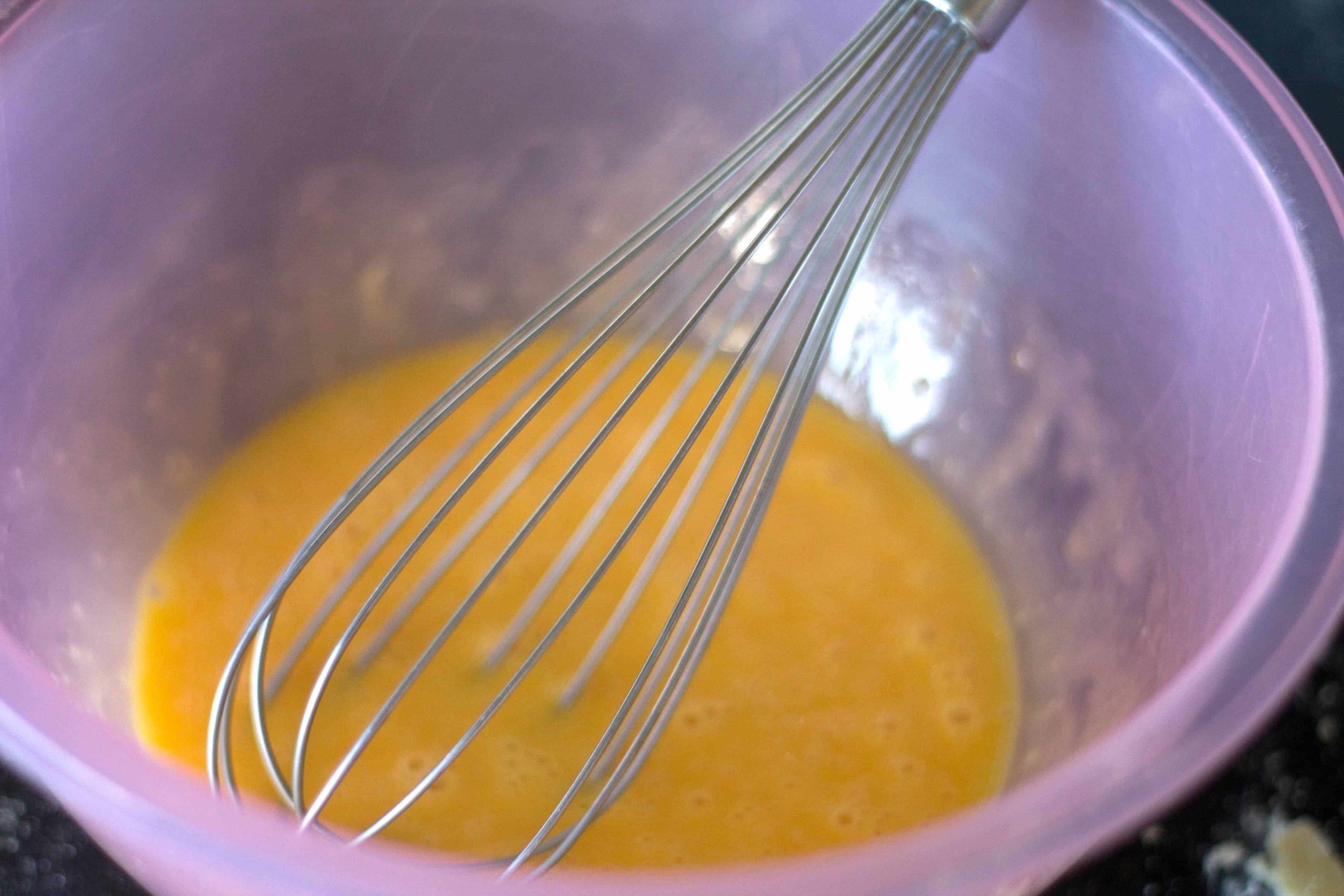 a pink plastic bowl with eggs being whisked with a metal egg whisk