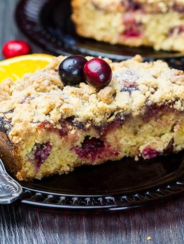 A piece of Cranberry Orange Crumb Cake with cranberries scattered around it in the table
