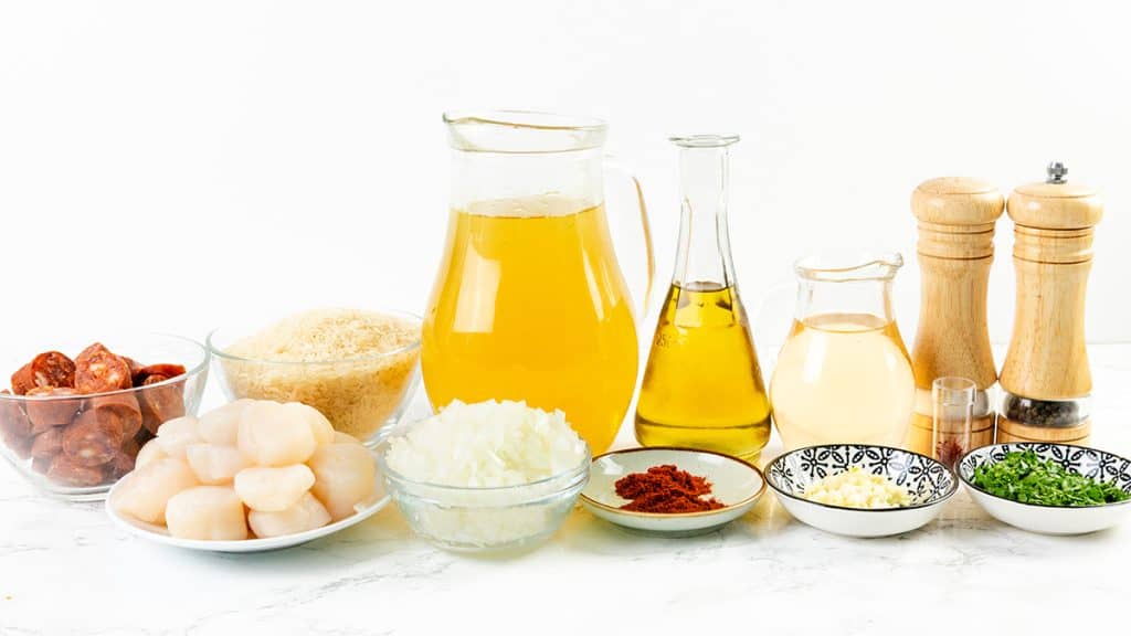 A variety of ingredients on a white surface—including chorizo slices, scallops, onions, rice, oil, broth, white wine, spices, garlic, chives, and seasonings—perfect for making Easy One Pot Scallops & Chorizo Paella.
