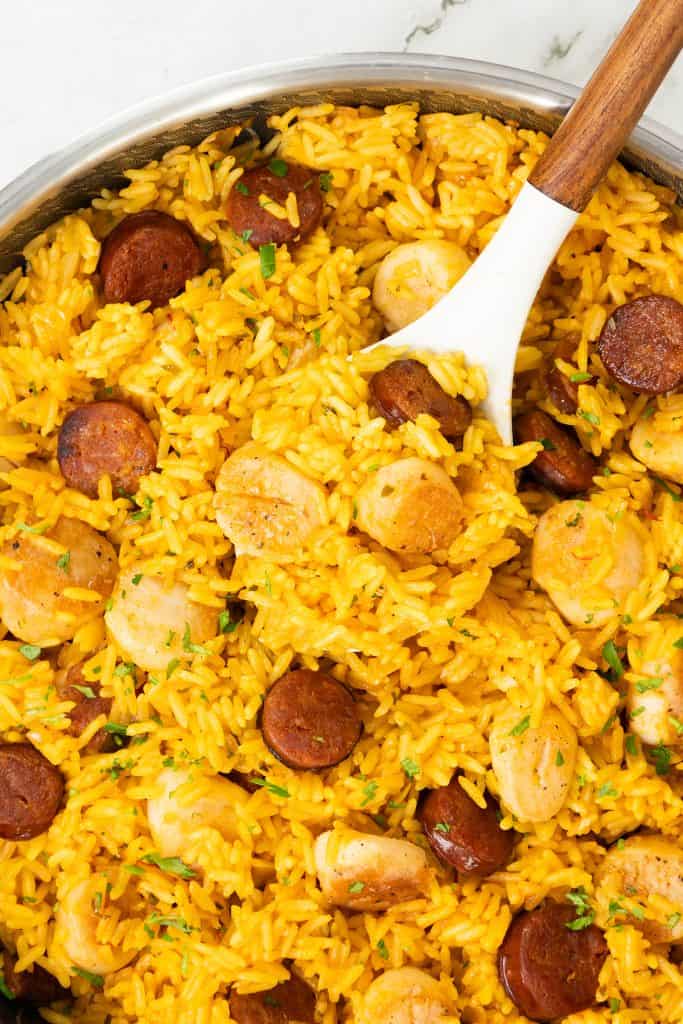 A pan filled with yellow rice, sliced sausage, and pieces of chicken being stirred with a white and wooden spoon—just like an Easy One Pot Scallops & Chorizo Paella. The dish is garnished with a sprinkle of fresh herbs.