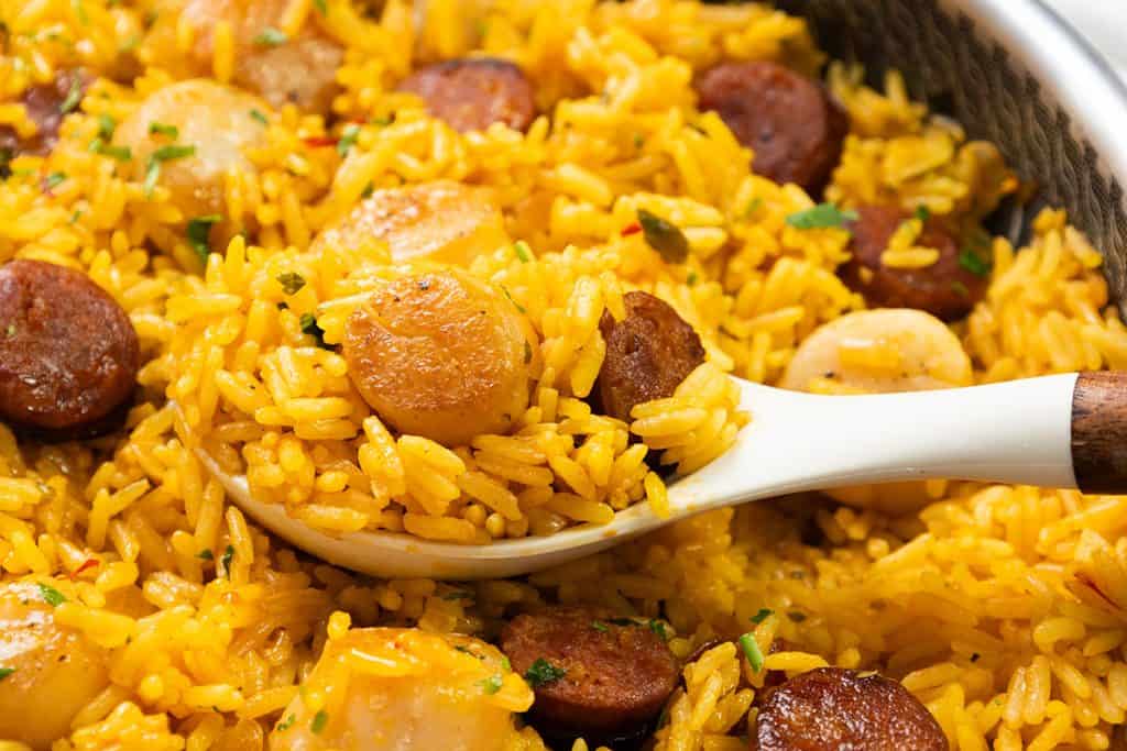 A close-up of Easy One Pot Scallops & Chorizo Paella, featuring yellow rice mixed with sliced sausage and scallops, served with a white ceramic spoon. The dish appears flavorful, garnished with chopped herbs.