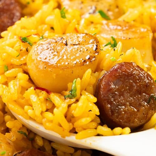 A close-up of Easy One Pot Scallops & Chorizo Paella—yellow rice with seared scallops and sausage slices, garnished with chopped herbs, served on a white spoon.