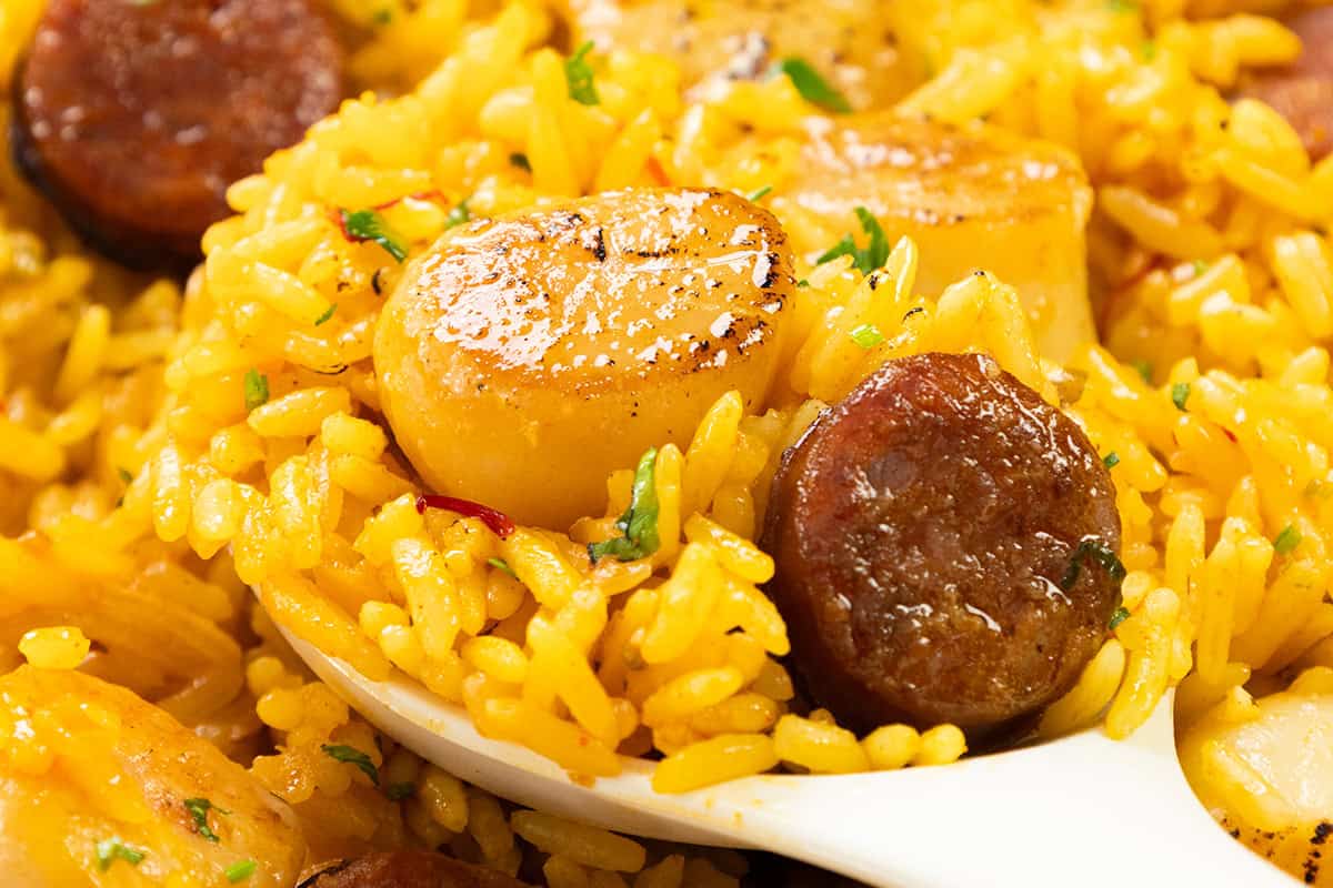 A close-up of Easy One Pot Scallops & Chorizo Paella—yellow rice with seared scallops and sausage slices, garnished with chopped herbs, served on a white spoon.