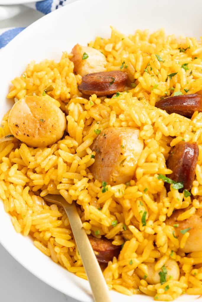 A close-up of a bowl filled with yellow rice, sliced sausage, and pieces of chicken, garnished with herbs. Reminiscent of an Easy One Pot Scallops & Chorizo Paella, a fork rests in the bowl, ready to eat.