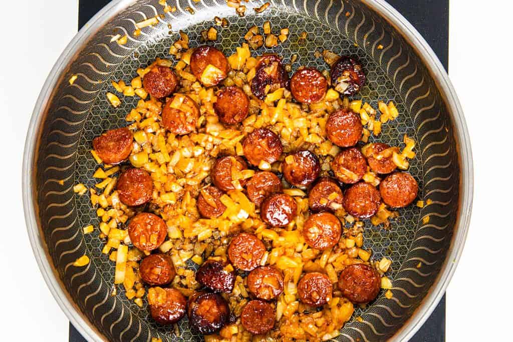 A frying pan with sliced chorizo sausage and chopped onions sizzling in oil, both browned to perfection—an aromatic start for an Easy One Pot Scallops & Chorizo Paella spread evenly across the patterned pan surface.