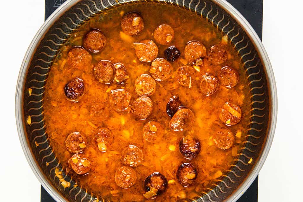 A frying pan filled with sliced sausage simmers in a reddish-brown sauce with chopped garlic and onions, resembling the rich flavors of an Easy One Pot Scallops & Chorizo Paella. The pan rests on a sleek black stovetop.