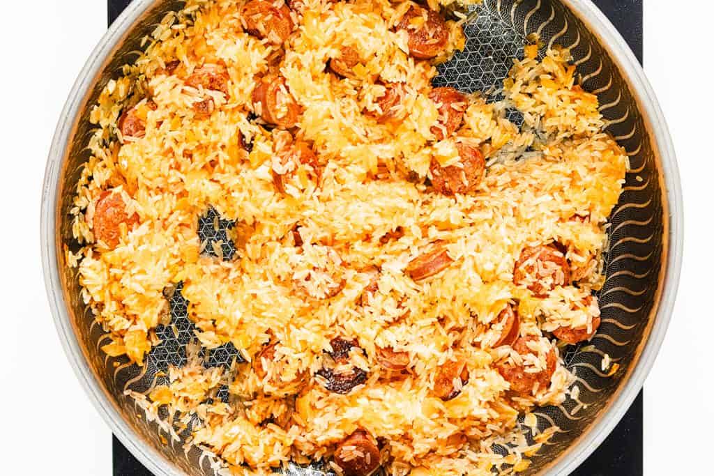 A close-up of a stainless steel pan filled with cooked yellow rice and sliced sausage pieces, inspired by Easy One Pot Scallops & Chorizo Paella, sitting on a stovetop. The rice is fluffy and mixed evenly with the sausage.
