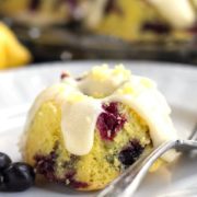 A close up of a lemon and blueberry cake with cream cheese icing drizzled over it, a fork and fresh blueberries
