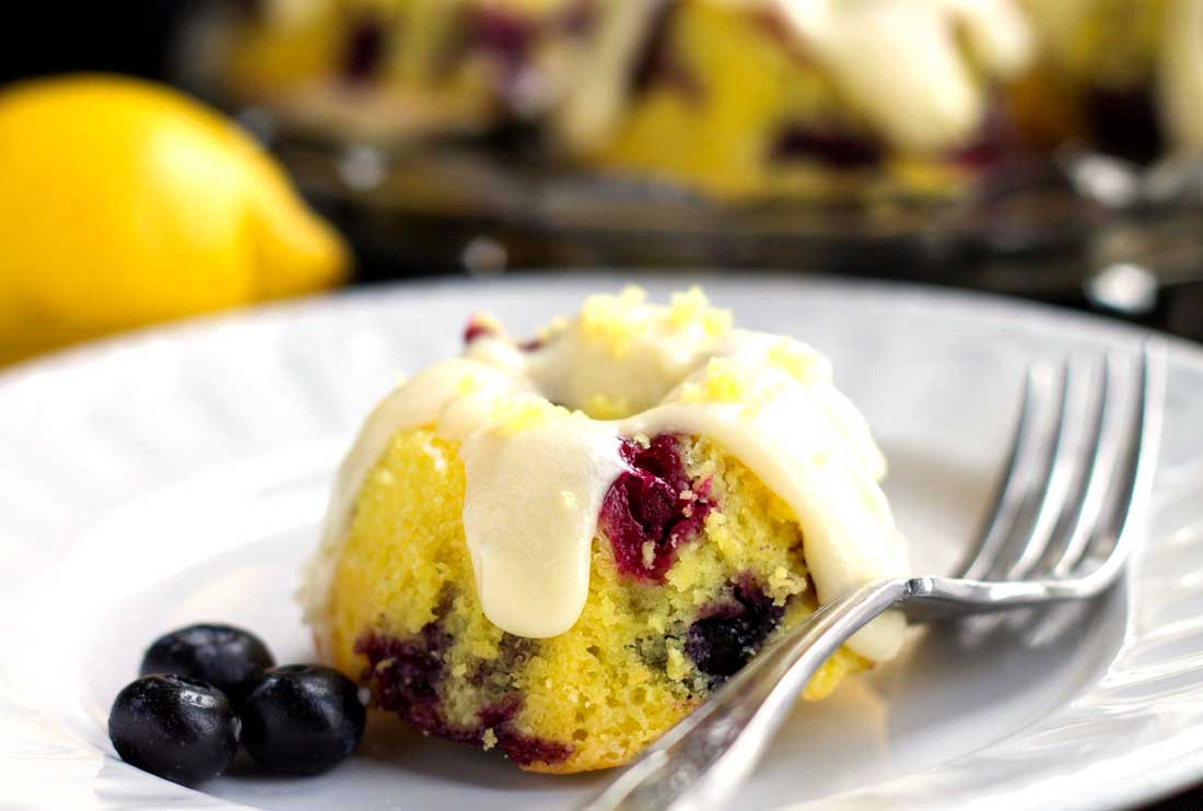 One frosted Lemon Blueberry Cake with blueberries and a fork next to it and more cakes and a lemon in the background.