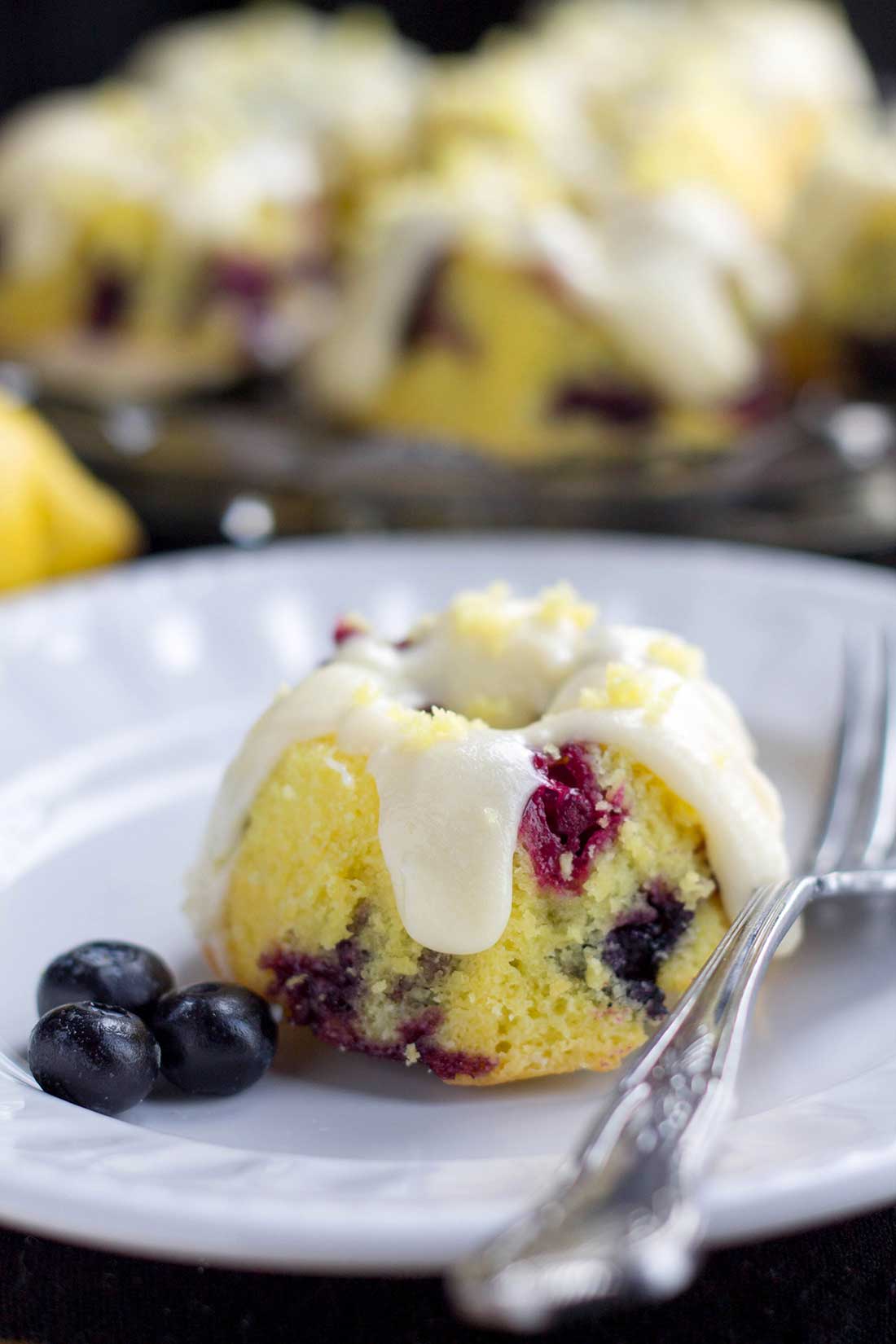 One frosted cake on a plate with blueberries and a fork on either side and more caked and a lemon in the background.