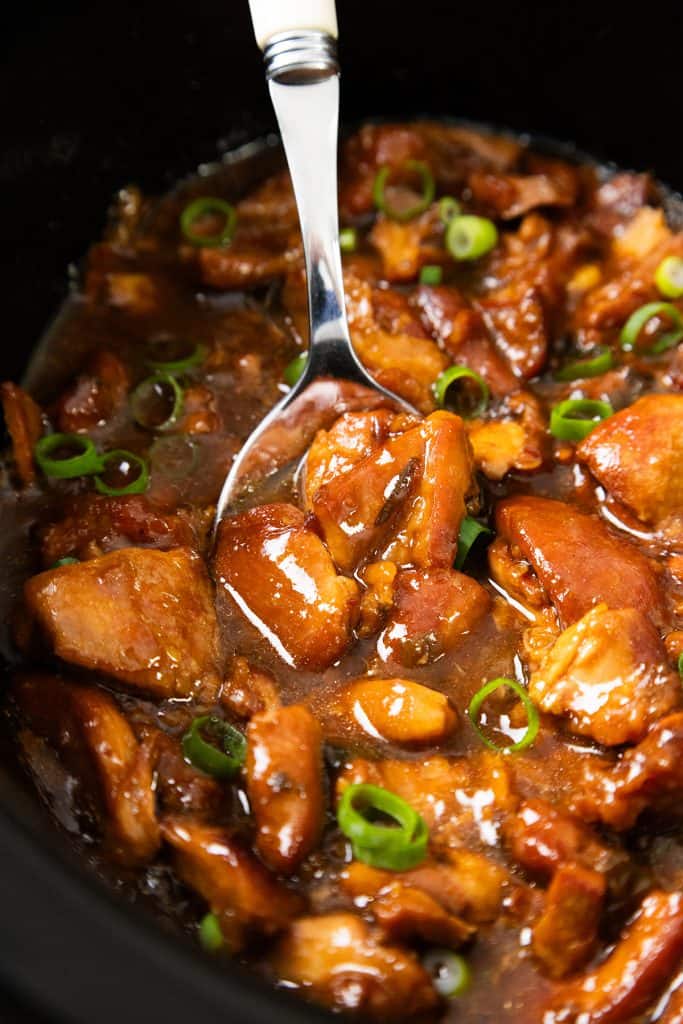 A close-up of Slow Cooker Honey Soy Chicken—tender pieces in a rich brown sauce, garnished with sliced green onions, with a metal serving spoon scooping some from a black pot.