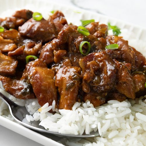 A plate of white rice topped with glazed, saucy Slow Cooker Honey Soy Chicken pieces and garnished with sliced green onions. A spoon rests on the side of the plate.