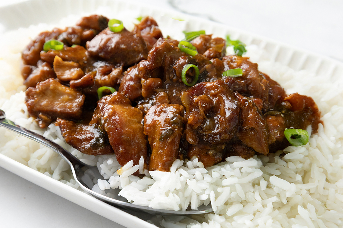A plate of white rice topped with glazed, saucy Slow Cooker Honey Soy Chicken pieces and garnished with sliced green onions. A spoon rests on the side of the plate.