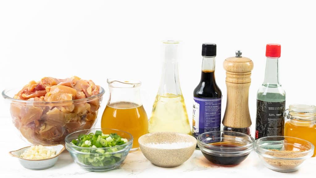 A variety of ingredients for Slow Cooker Honey Soy Chicken on a white surface, including a large bowl of diced raw chicken, chopped green onions, minced garlic, sauces, seasonings, pepper grinder, and bottles of soy sauce and vinegar.
