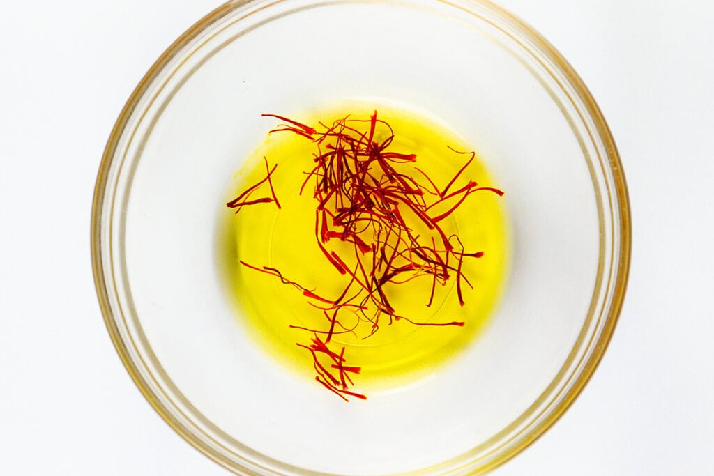 A glass bowl filled with yellow liquid, likely oil, with red saffron threads floating on top, evokes the vibrant colors of Tagliarini with Saffron Cream Sauce, viewed from above against a white background.