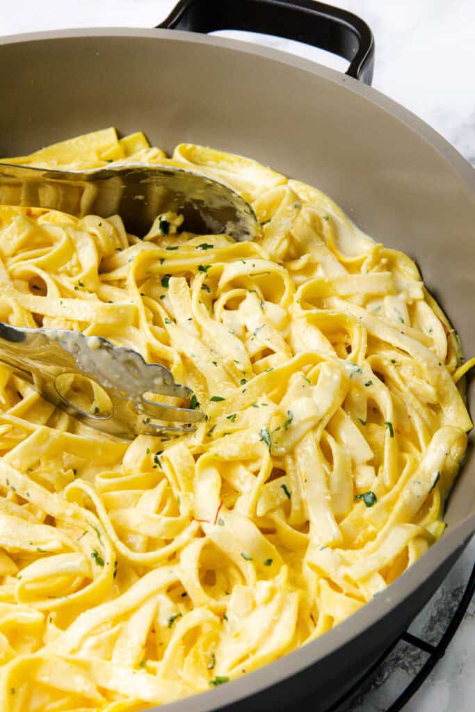 A pan filled with creamy Tagliarini with Saffron Cream Sauce pasta, garnished with chopped herbs, with metal tongs resting on top, ready for serving.