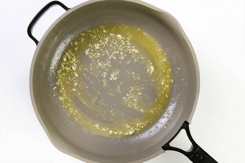 A gray skillet with melted butter and minced garlic sizzling, the perfect start for Tagliarini with Saffron Cream Sauce, viewed from above on a white background.