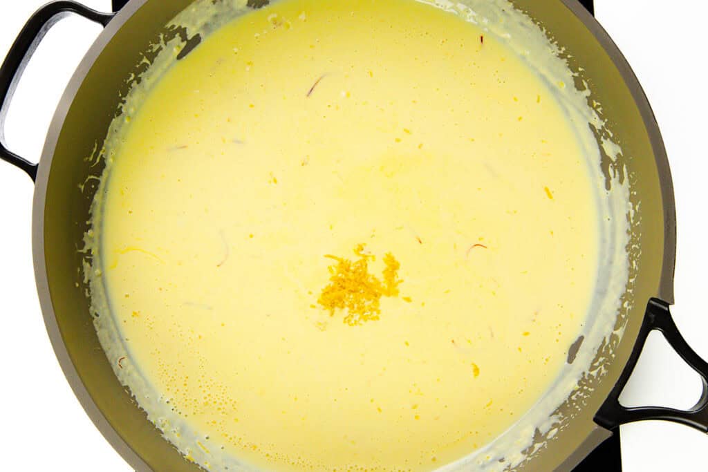 A close-up of a pot filled with creamy yellow Tagliarini with Saffron Cream Sauce, topped with a small pile of lemon zest in the center. The pot has black handles and sits on a white surface.