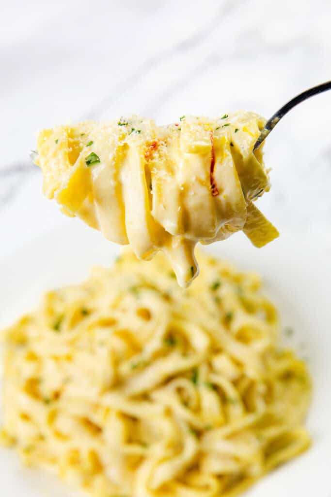 A close-up of a fork twirling creamy Tagliarini with Saffron Cream Sauce above a plateful of pasta, garnished with grated cheese and herbs, on a white background.