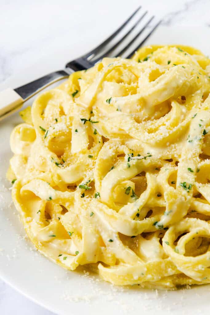 A plate of creamy Tagliarini with Saffron Cream Sauce, garnished with grated parmesan cheese and chopped parsley, with a fork and knife placed beside it on a white surface.