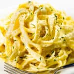 A close-up of creamy Tagliarini with Saffron Cream Sauce, topped with grated Parmesan cheese and chopped parsley, served on a white plate with a fork.
