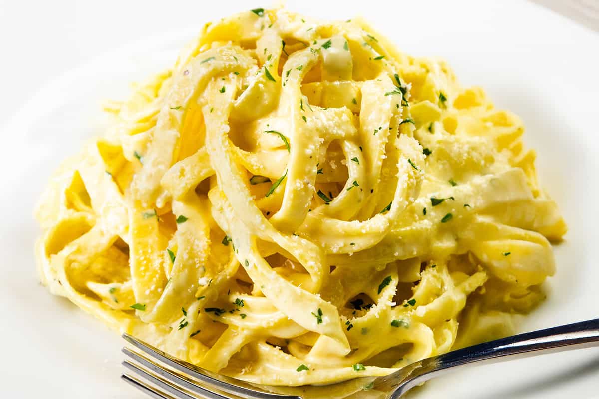 A close-up of creamy Tagliarini with Saffron Cream Sauce, topped with grated Parmesan cheese and chopped parsley, served on a white plate with a fork.