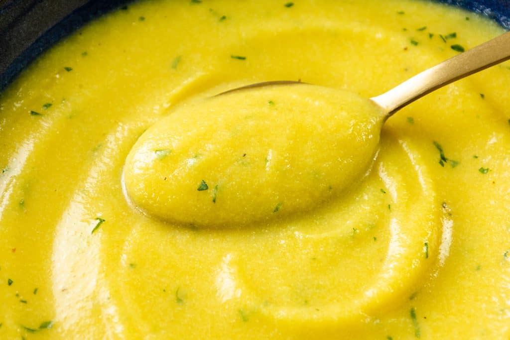 A close-up of a spoonful of low fat creamy cauliflower saffron soup with green herbs, held above a bowl filled with the same smooth, thick soup.