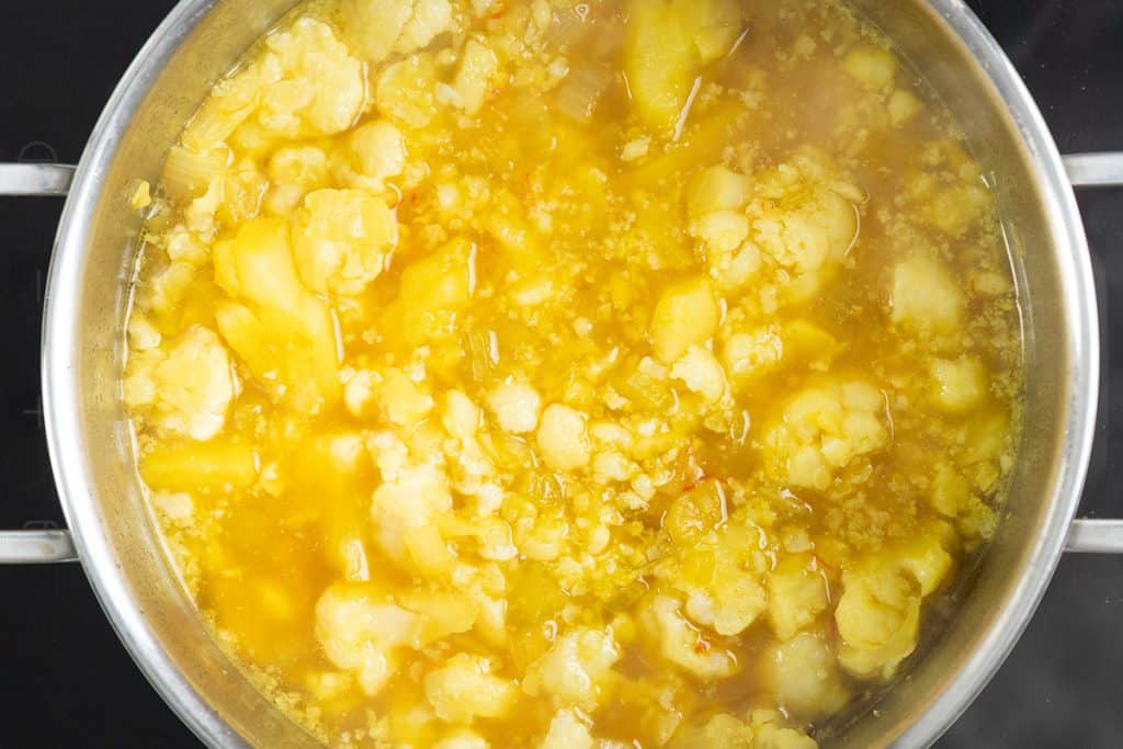 A pot of Low Fat Creamy Cauliflower Saffron Soup with visible chunks of cauliflower and potatoes simmers in golden broth on the stove.