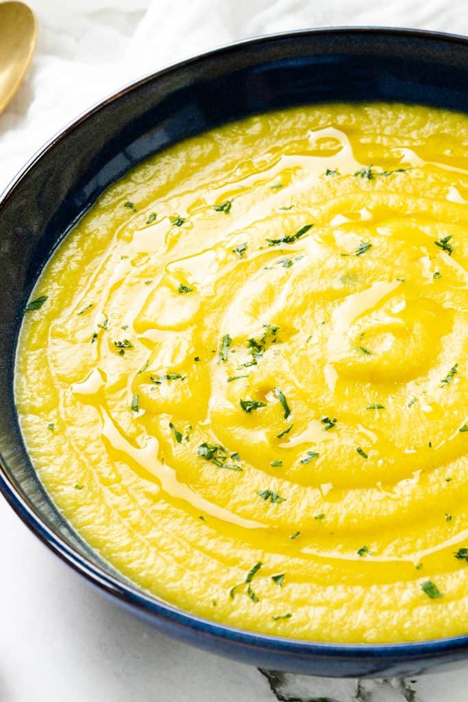 A bowl of Low Fat Creamy Cauliflower Saffron Soup, with its golden hue, is garnished with chopped herbs and a drizzle of olive oil, served in a dark blue bowl on a marble surface.