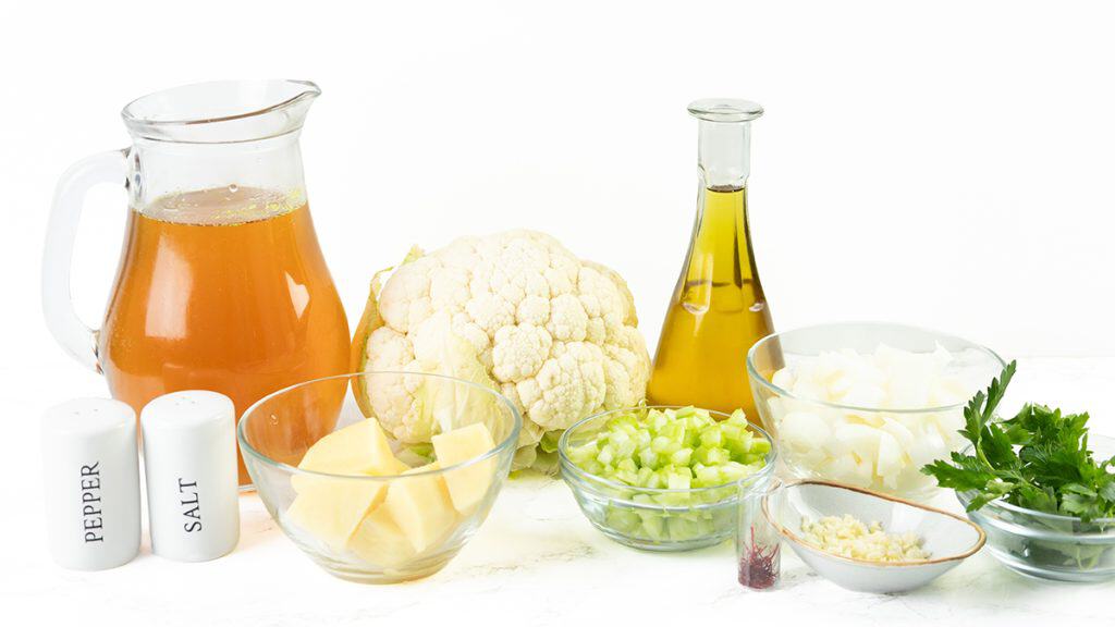 A pitcher of broth, a head of cauliflower, a bottle of oil, bowls of chopped onion, celery, potato, garlic, parsley, and seasonings are arranged on a white surface&mdash;everything you need to make Low Fat Creamy Cauliflower Saffron Soup.