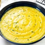 A bowl of Low Fat Creamy Cauliflower Saffron Soup, its yellow hue rich and inviting, is garnished with fresh herbs and served alongside grilled bread slices. Gold spoons and a sprinkle of chopped herbs rest nearby on a pristine white surface.