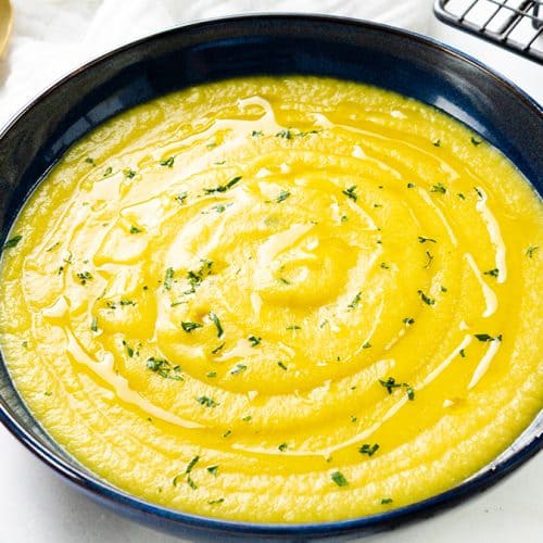 A bowl of Low Fat Creamy Cauliflower Saffron Soup, its yellow hue rich and inviting, is garnished with fresh herbs and served alongside grilled bread slices. Gold spoons and a sprinkle of chopped herbs rest nearby on a pristine white surface.