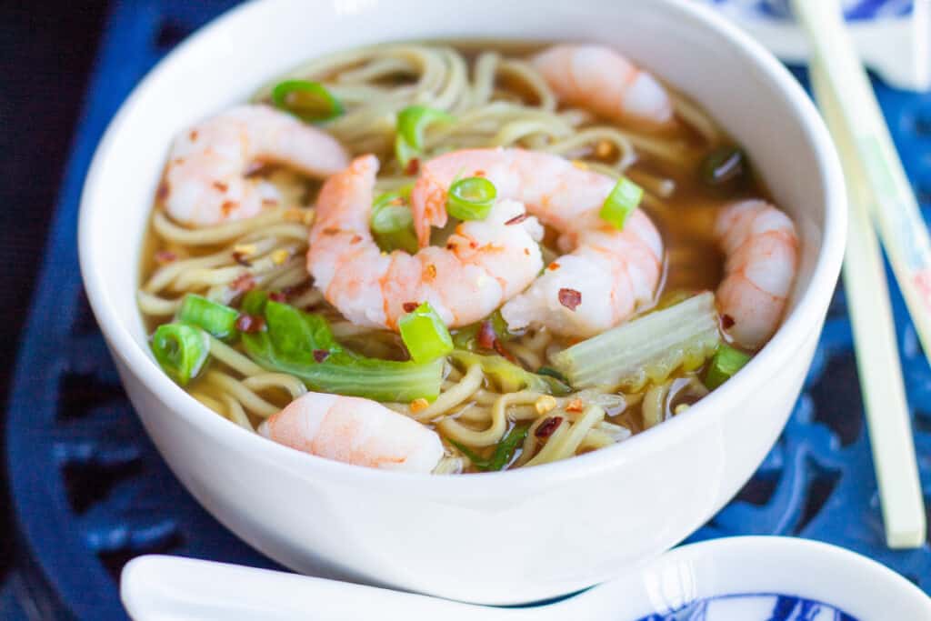 A close up of a bowl of Chinese Shrimp noodle soup with scattered chili flakes and sliced green onions