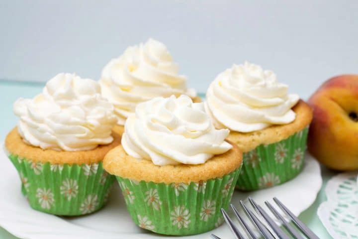 A serving plate filled with Peaches and Cream Cupcakes topped with whipped cream frosting with peaches in the background