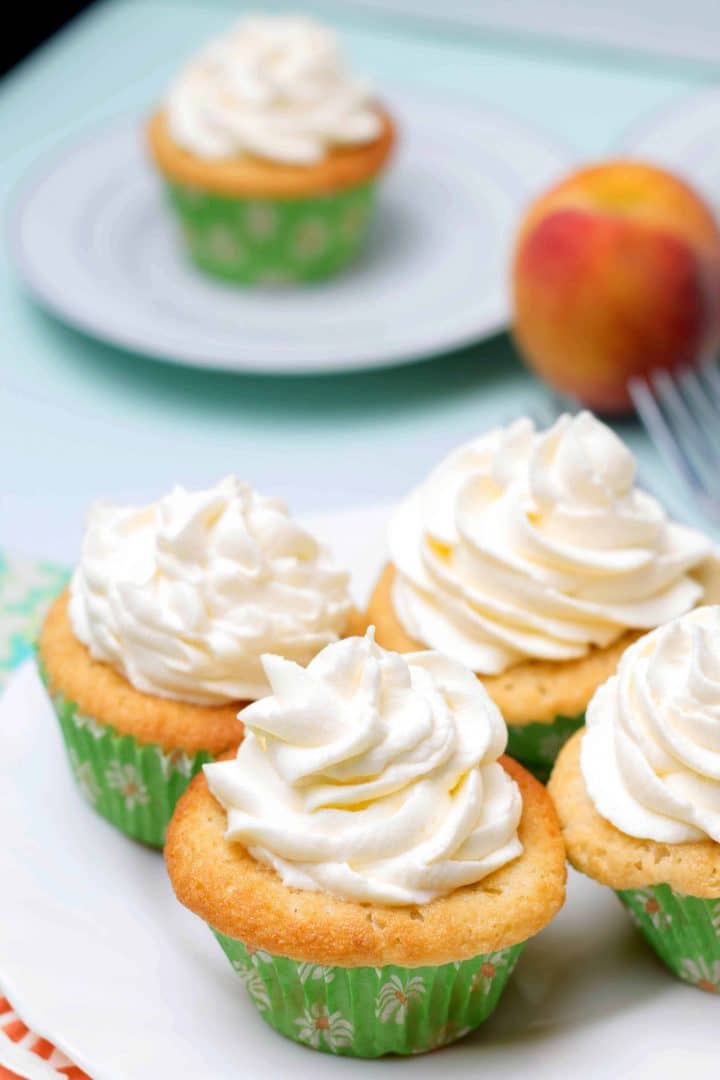 Four Peaches and Cream Cupcake on a serving plate piled high with whipped cream frosting with a peach in the background