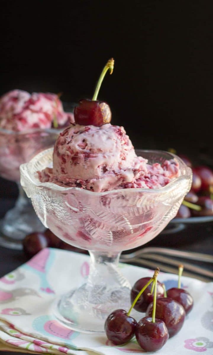 Cherry ice cream in a pretty glass bowl topped with a fresh cherry with more ice cream and cherries in the background.