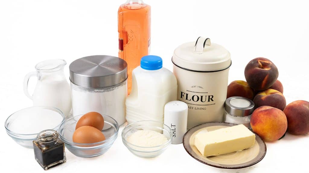 Baking ingredients including flour, sugar, eggs, milk, butter, vanilla, salt, oil, baking powder, and several fresh peaches are arranged on a white surface—perfect for making delightful Peaches and Cream Cupcakes.