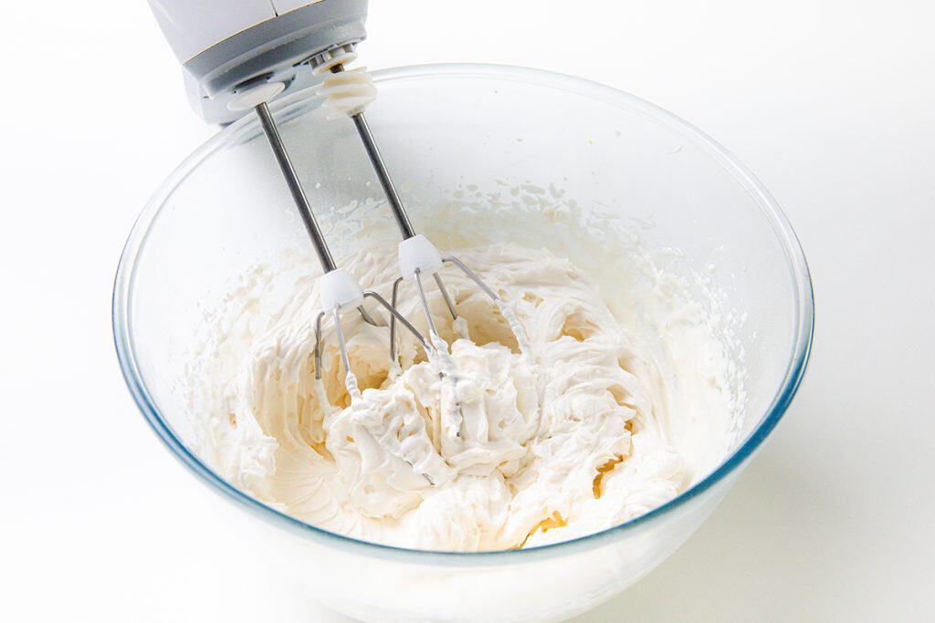 A hand mixer with metal beaters is mixing fluffy whipped cream for Peaches and Cream Cupcakes in a clear glass bowl on a white surface.