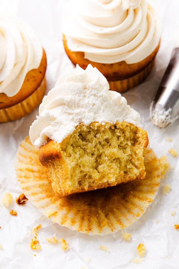 A Peaches and Cream Cupcake with white frosting sits on a crinkled wrapper, showing a large bite taken out. Two whole frosted cupcakes and a metal piping tip are in the background. Crumbs are scattered around.