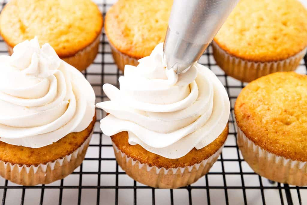 A piping bag adds swirls of white frosting onto Peaches and Cream Cupcakes lined up on a black wire cooling rack. Some cupcakes are already frosted, while others remain plain.