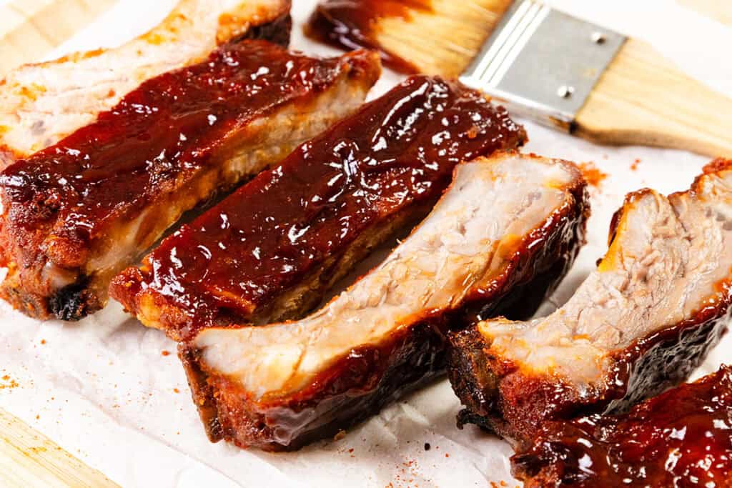 Baked or Barbecued Sticky Glazed Ribs coated in a thick, glossy sauce are arranged on parchment paper, with a basting brush in the background. The ribs are tender, juicy, and slightly charred along the edges.