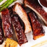 Baked or Barbecued Sticky Glazed Ribs are arranged on a surface with asparagus spears, grilled corn, cherry tomatoes, and a cup of barbecue sauce visible in the corner.