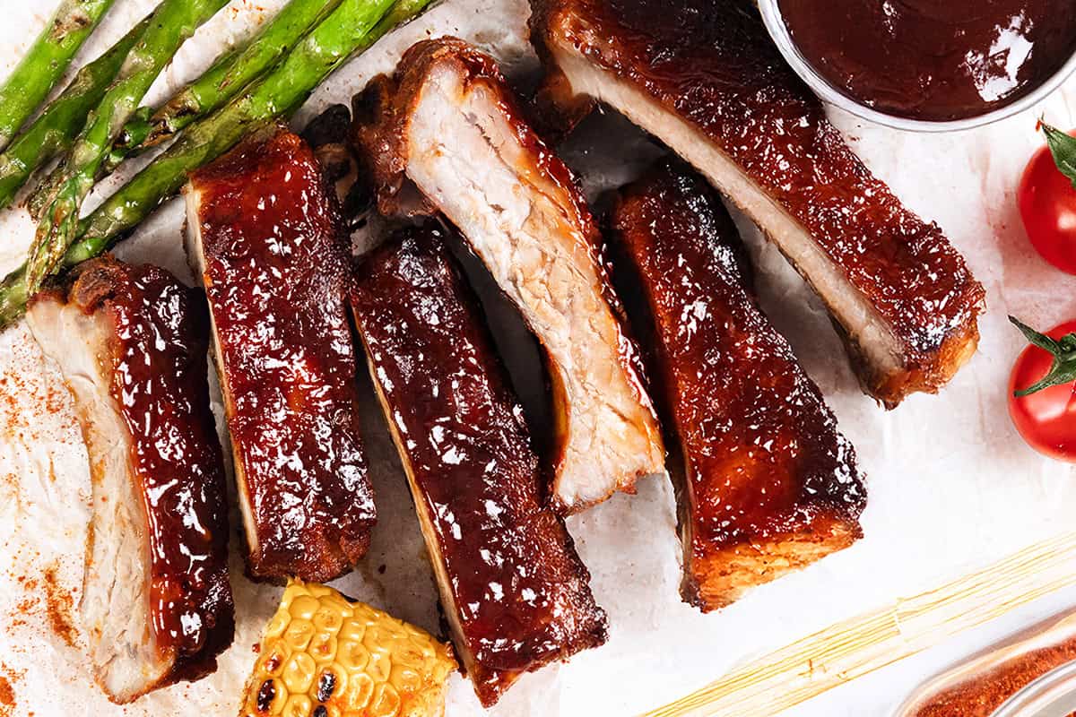 Baked or Barbecued Sticky Glazed Ribs are arranged on a surface with asparagus spears, grilled corn, cherry tomatoes, and a cup of barbecue sauce visible in the corner.
