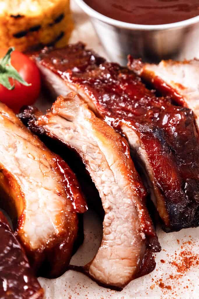 Close-up of baked or barbecued sticky glazed ribs with a shiny, caramelized crust, served next to a cherry tomato, a slice of cornbread, and a metal cup of barbecue sauce in the background.