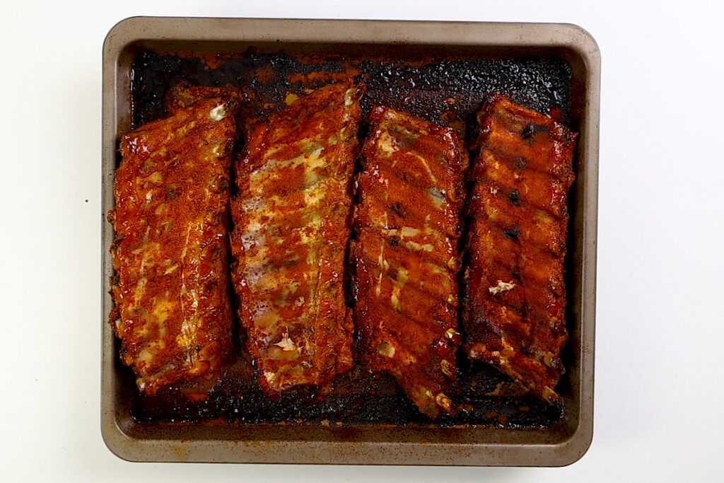 A rectangular baking tray with four cooked racks of Baked Sticky Glazed Ribs, coated in a reddish-brown sauce and arranged side by side on a white background.