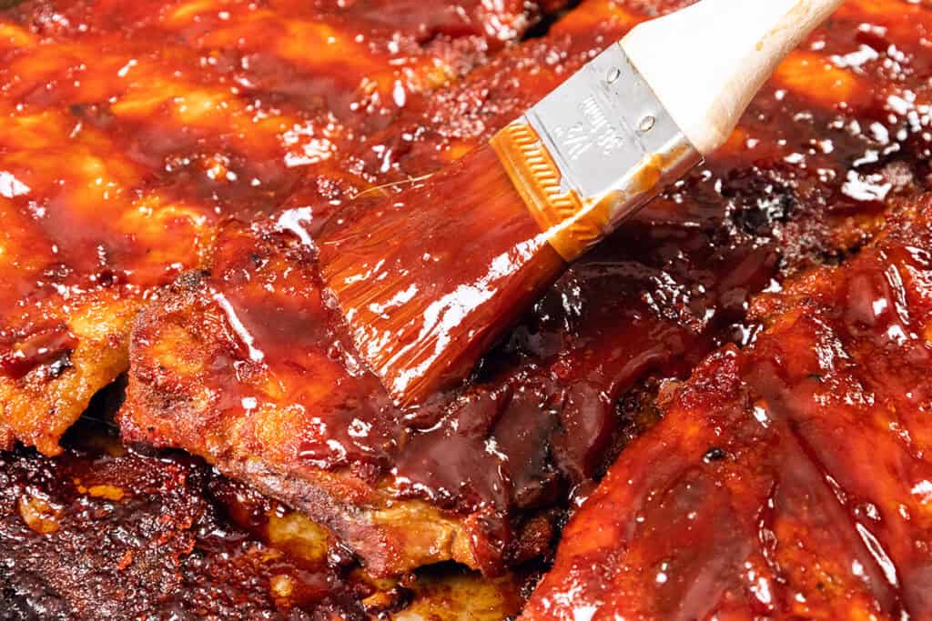 A close-up of Baked or Barbecued Sticky Glazed Ribs being coated with a thick layer of glossy barbecue sauce using a basting brush. The ribs appear juicy and caramelized, with a rich reddish-brown color.
