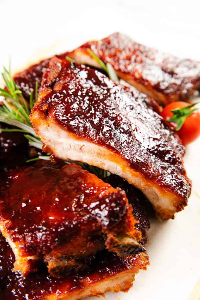 Close-up of Baked or Barbecued Sticky Glazed Ribs stacked on a plate, garnished with fresh rosemary and cherry tomatoes. The ribs are coated in a shiny, rich sauce.
