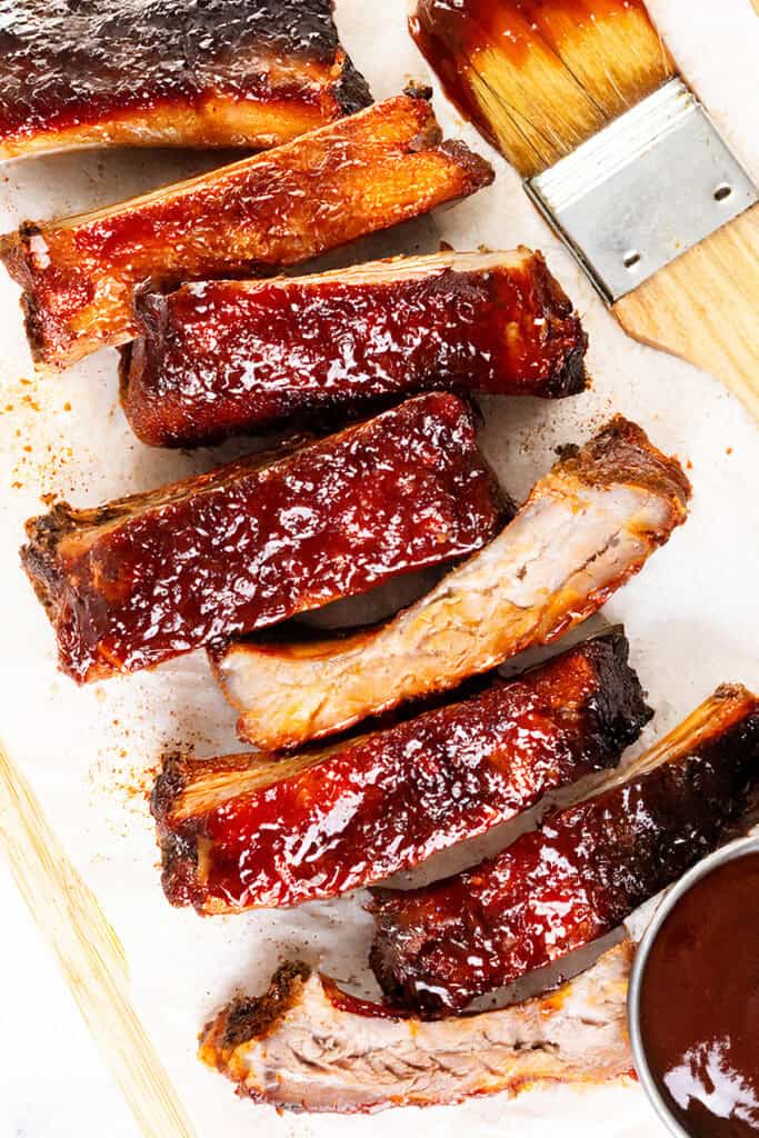 Baked or Barbecued Sticky Glazed Ribs are coated in glossy sauce and arranged on parchment paper, with a basting brush and a small bowl of barbecue sauce nearby. The ribs appear tender with a caramelized surface.