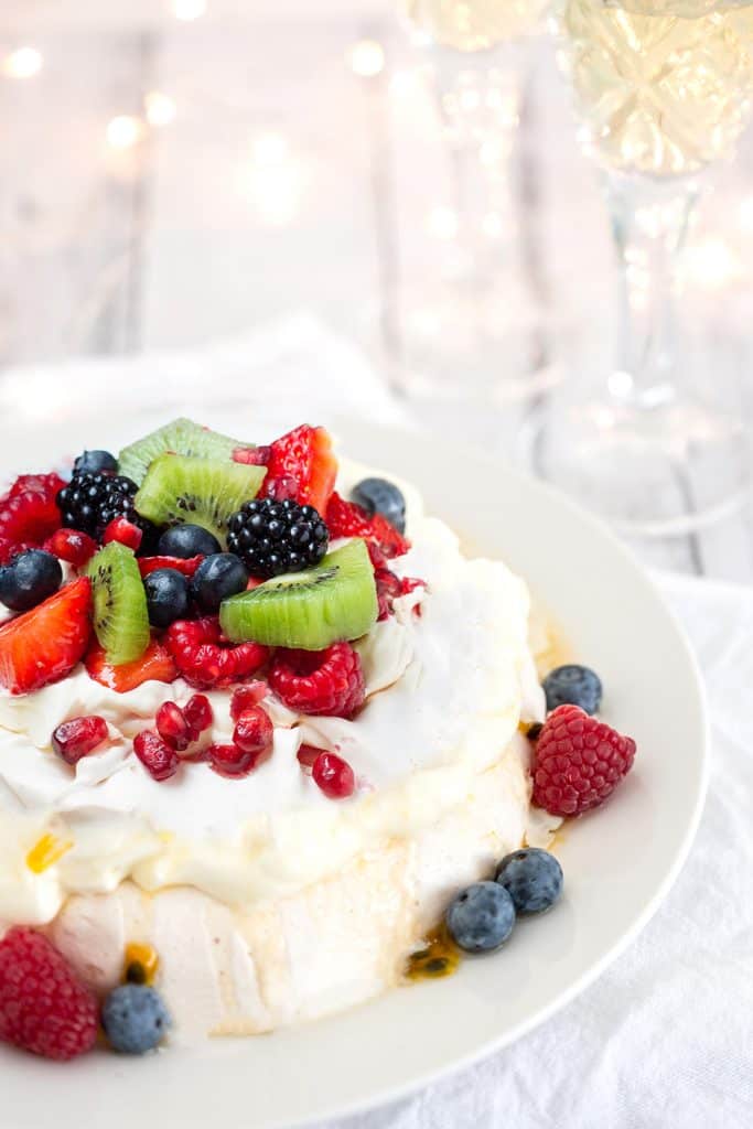 Pavlova with strawberries, blueberries, raspberries and passionfruit with Christmas lights and sparkling wine in the background to celebrate the holidays in Australia